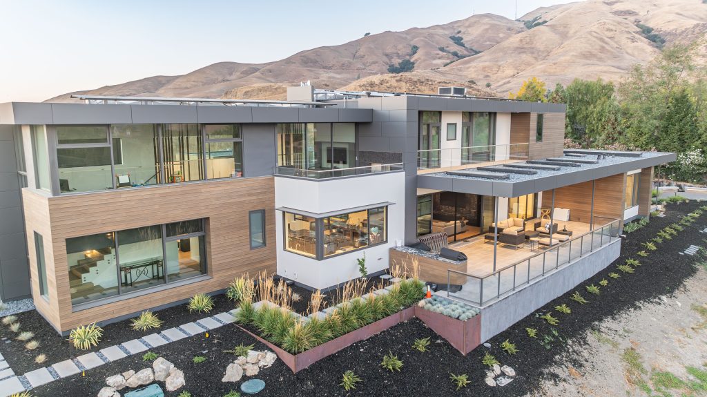 A luxurious custom-built home by JCD Builders Inc., featuring a sleek modern design with large glass windows, a spacious outdoor living area, and a scenic mountain backdrop. The house combines wood, glass, and metal elements, showcasing contemporary architecture that blends seamlessly with nature.